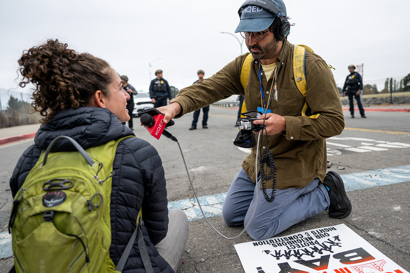 Reporter Azul Dahlstrom-Eckman wears a cap labeled “KQED,” over-ear headphones, glasses, and a yellow backpack kneels on a paved street while conducting an interview with a protester  in front of the entrance to a U.S. Coast Guard base in Oakland on Oct. 23, 2025. He holds a red KQED-branded microphone toward the female protester seated on the ground, who wears a dark puffer jacket and a bright green backpack. Dahlstrom-Eckman also holds a small audio recorder with cables attached. In the background, several police officers stand spaced out across the road, slightly out of focus. A protest sign with partially visible text lies on the ground in the foreground near the reporter’s knee. The sky is overcast, and the overall scene suggests an on-location interview during a public demonstration or street closure.