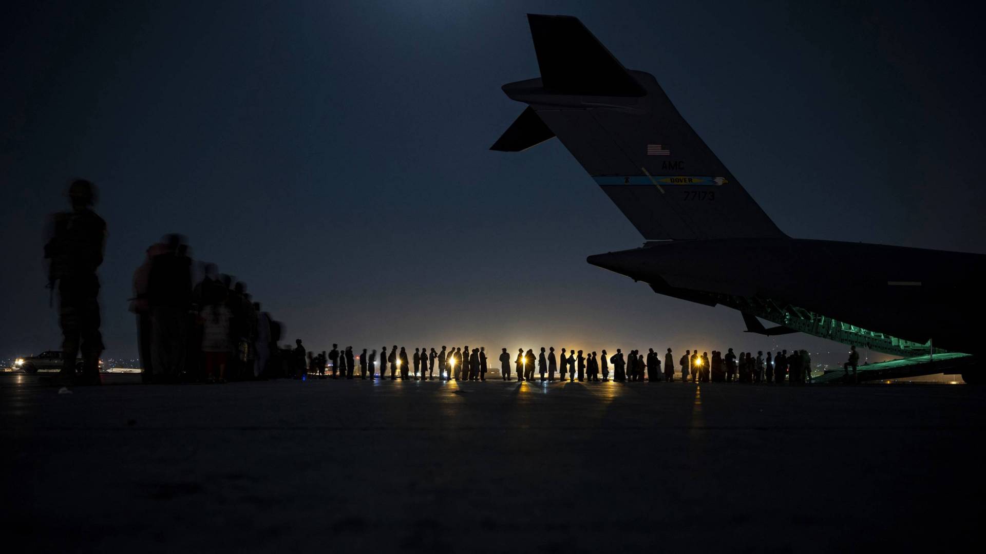 line of evacuees at night wait to board a plane, backlight by far off light