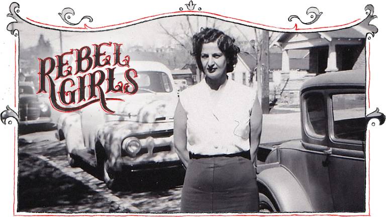 A black and white image of a 1940s-era woman standing near trucks and cars.