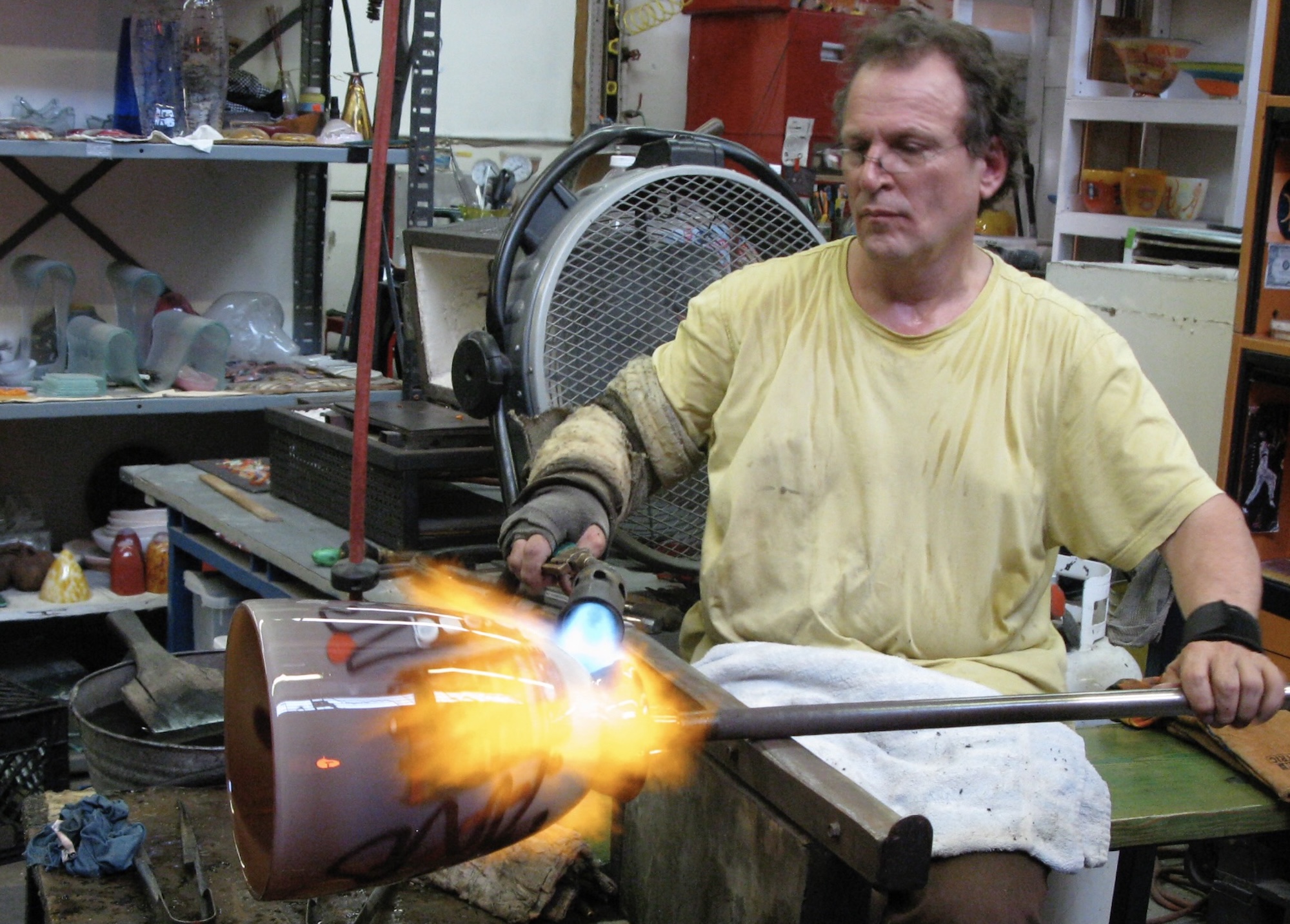 A middle-aged white man wearing glasses uses a blow torch on an ornate glass ornament attached to the end of a rod. He is seated inside a studio lined with shelves covered with other glass objects. 