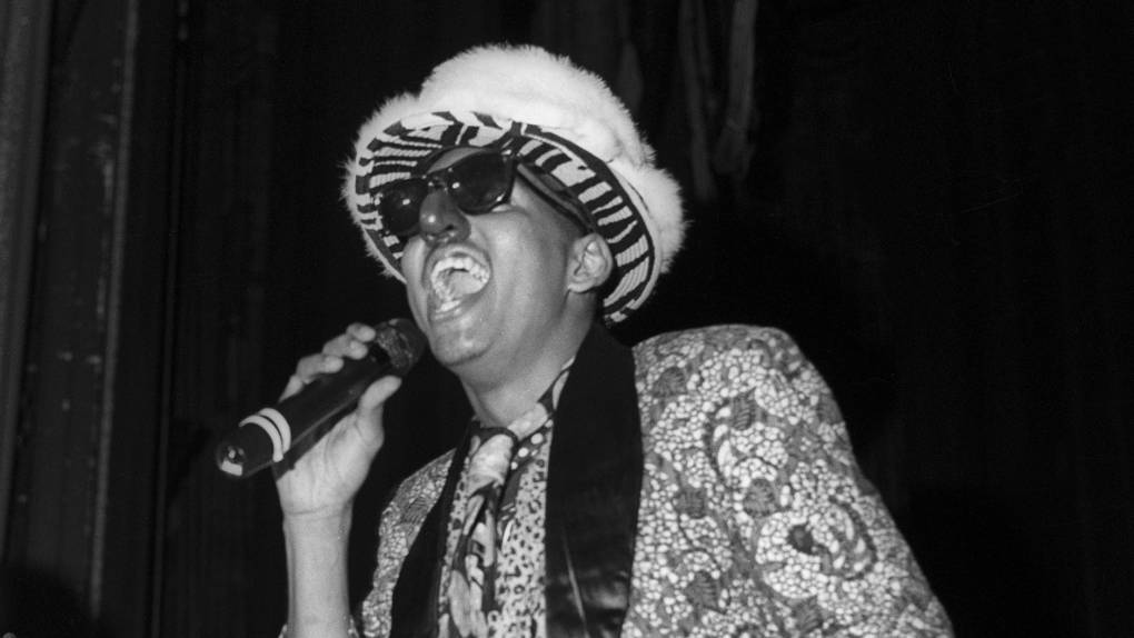 A black-and-white photo of Shock G performing, wearing a fur hat over a zebra-print hat, a patterned blazer and a fake nose. He holds a champagne bottle as he raps into the mic.