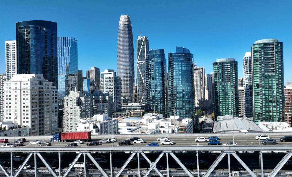 A city skyline with high-rises and a blue sky, cars driving in the foreground on a bridge.