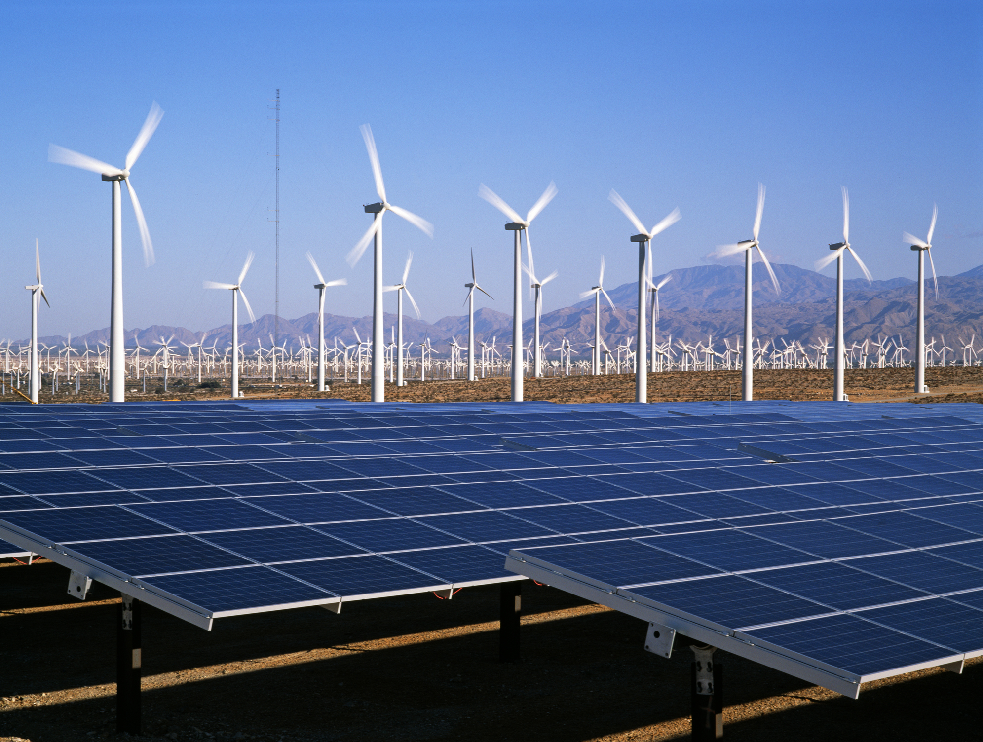 Solar panels and wind turbines in desert with mountains in the distance.