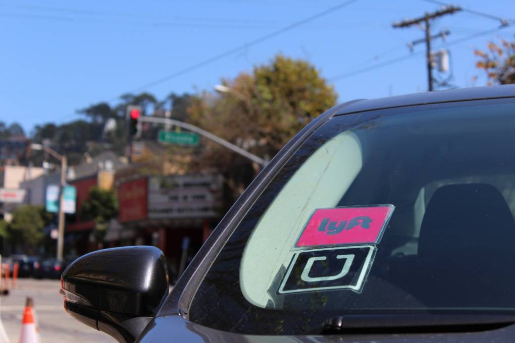 A close-up shot of a black vehicle with a pink Lyft sticker and a black-and-white Uber sticker on the left side of its windshield. The vehicle sits idle, waiting to pick up a customer.