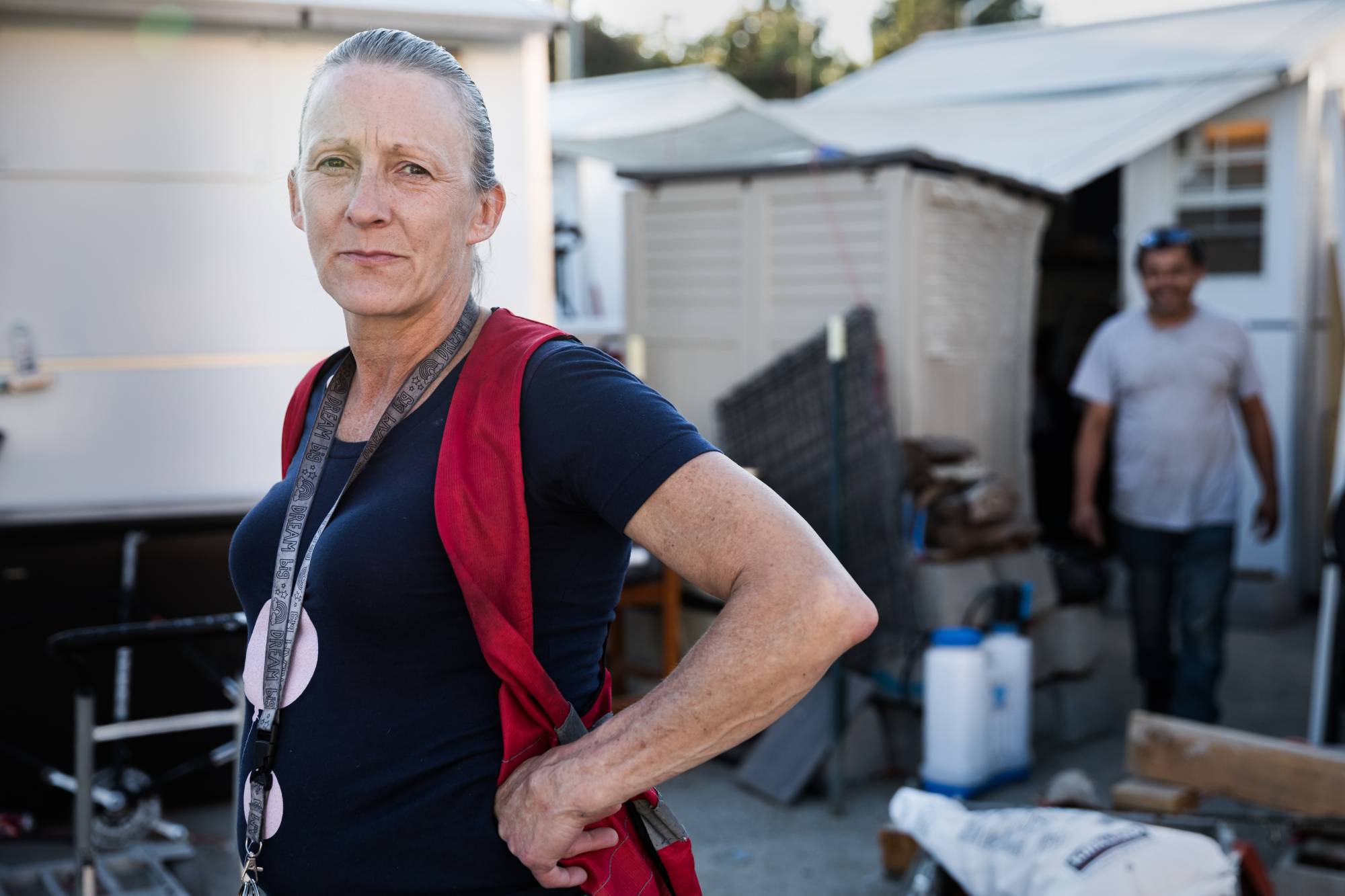 Tammy, a former resident of Oakland's East 12th Street Parcel, stands outdoors in the parcel, facing the camera with a steady, confident expression and one hand resting on her hip. She wears a dark short-sleeve shirt and a red vest over her shoulders, along with a lanyard hanging around her neck. Her hair is pulled back, and natural light highlights her face.

Behind her are small structures or sheds, a canopy or tarp, and various items such as containers, tools, and stacked materials. A second person is visible in the background, slightly out of focus, walking through the space. The setting suggests a lived-in, transitional outdoor environment, and her posture conveys resilience and self-possession.