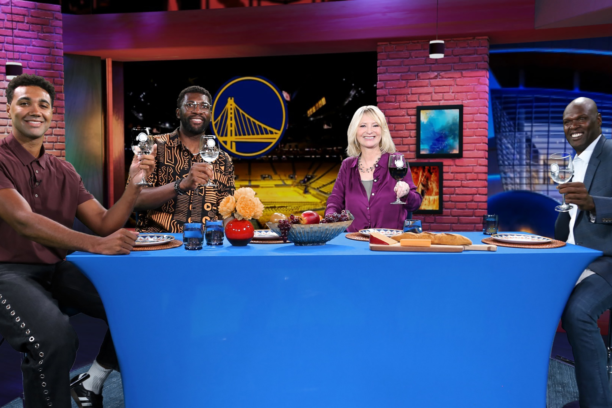 Three program guests and Check, Please! Bay Area host Leslie Sbrocco sit around a blue table on a studio set, smiling and raising wine glasses in a toast. Behind them is the Golden State Warriors logo inside a basketball arena backdrop. From left to right are  former Golden State Warriors Trayce Jackson-Davis, Festus Ezeli, Check, Please! Bay Area host Leslie Sbrocco, and former Golden State Warrior Adonal Foyle. The table is set with bread, cheese, fruit, and flowers, creating a casual, celebratory atmosphere.